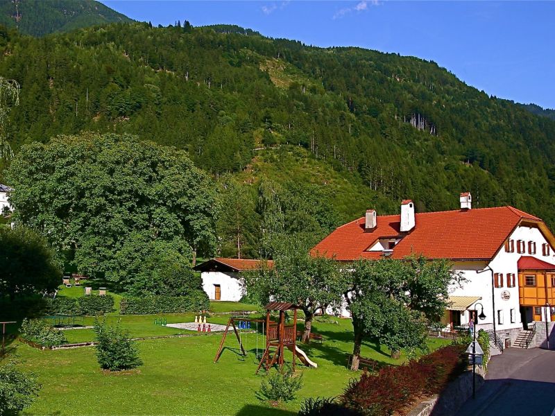 Außenansicht des Spiele- und Buchhotels Tschitscher mit rotem Dach, Garten und kleinem Spielplatz, im Grünen gelegen