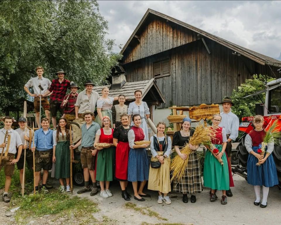 Eine Gruppe von Menschen in traditioneller Kleidung steht vor einer rustikalen Scheune und einem Traktor.