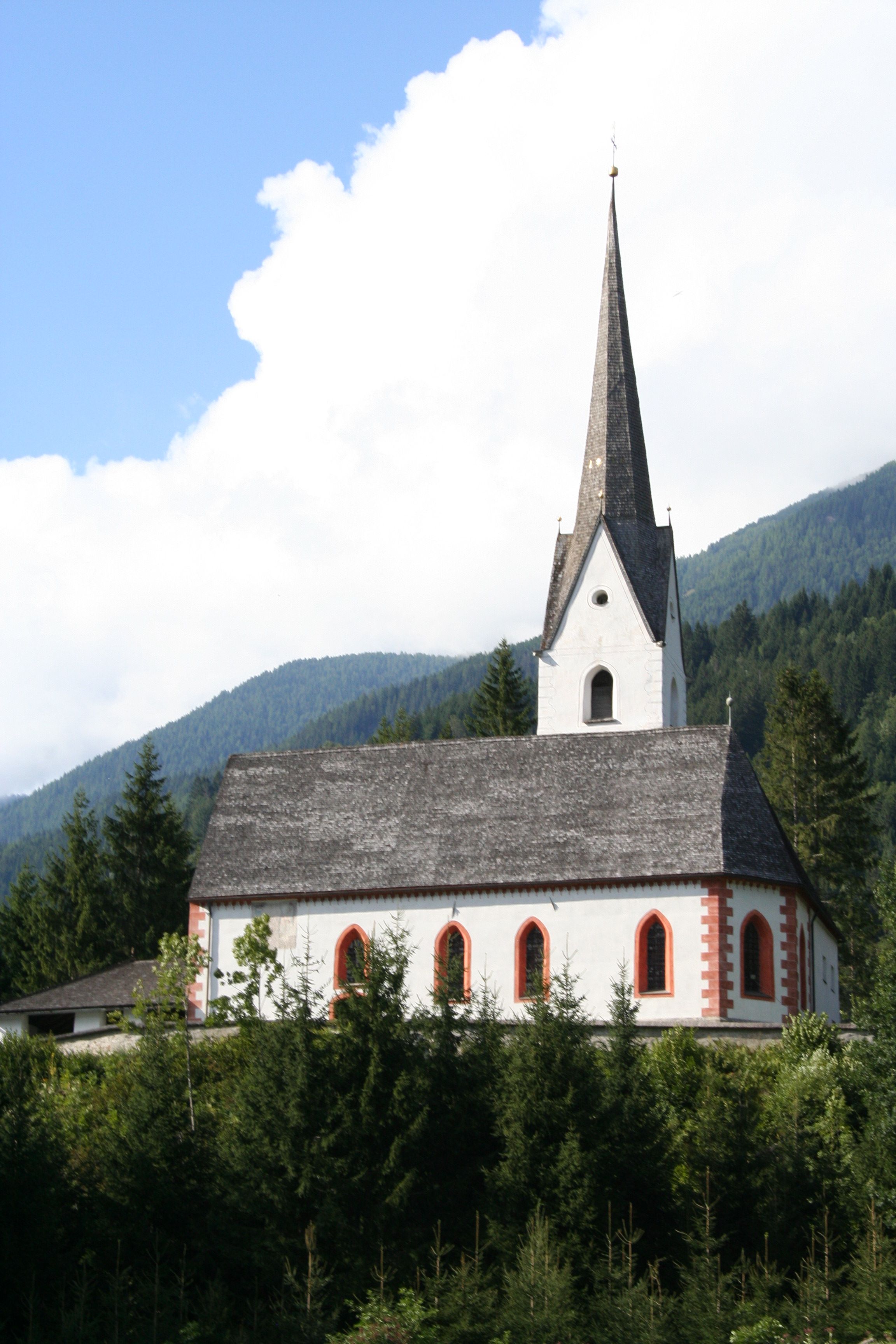 Eine bezaubernde weiße Kirche mit einem hohen Kirchturm, umgeben von üppigem Grün und einem klaren blauen Himmel, vor einer Bergkulisse.