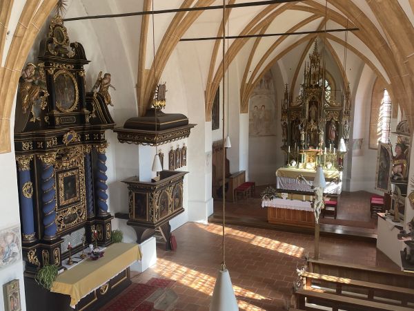 Blick in das Innere der Chrysanthner Pfarrkirche mit gotischem Gewölbe, Holzbänken und einem reich verzierten Altar im Sonnenlicht. 
