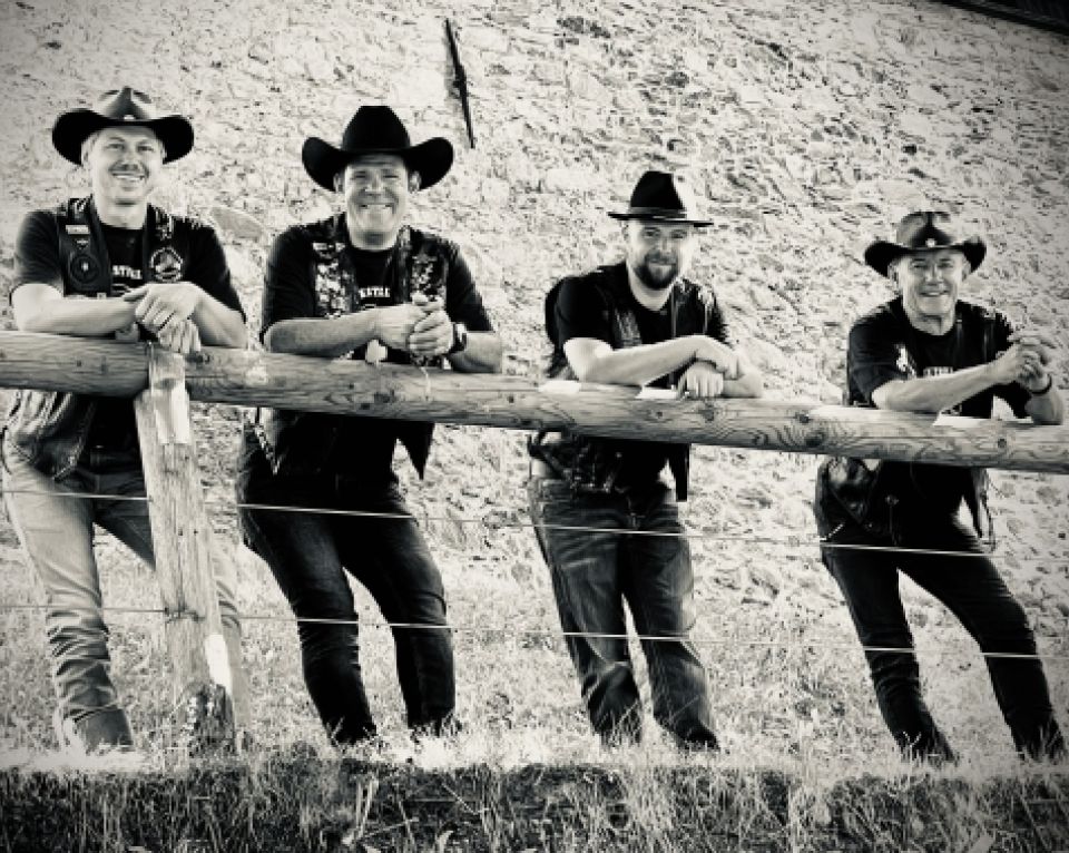 Eine Gruppe von vier Personen mit schwarzen Cowboyhüten, gekleidet in einheitlicher schwarzer Kleidung, lehnt lässig an einem Holzzaun