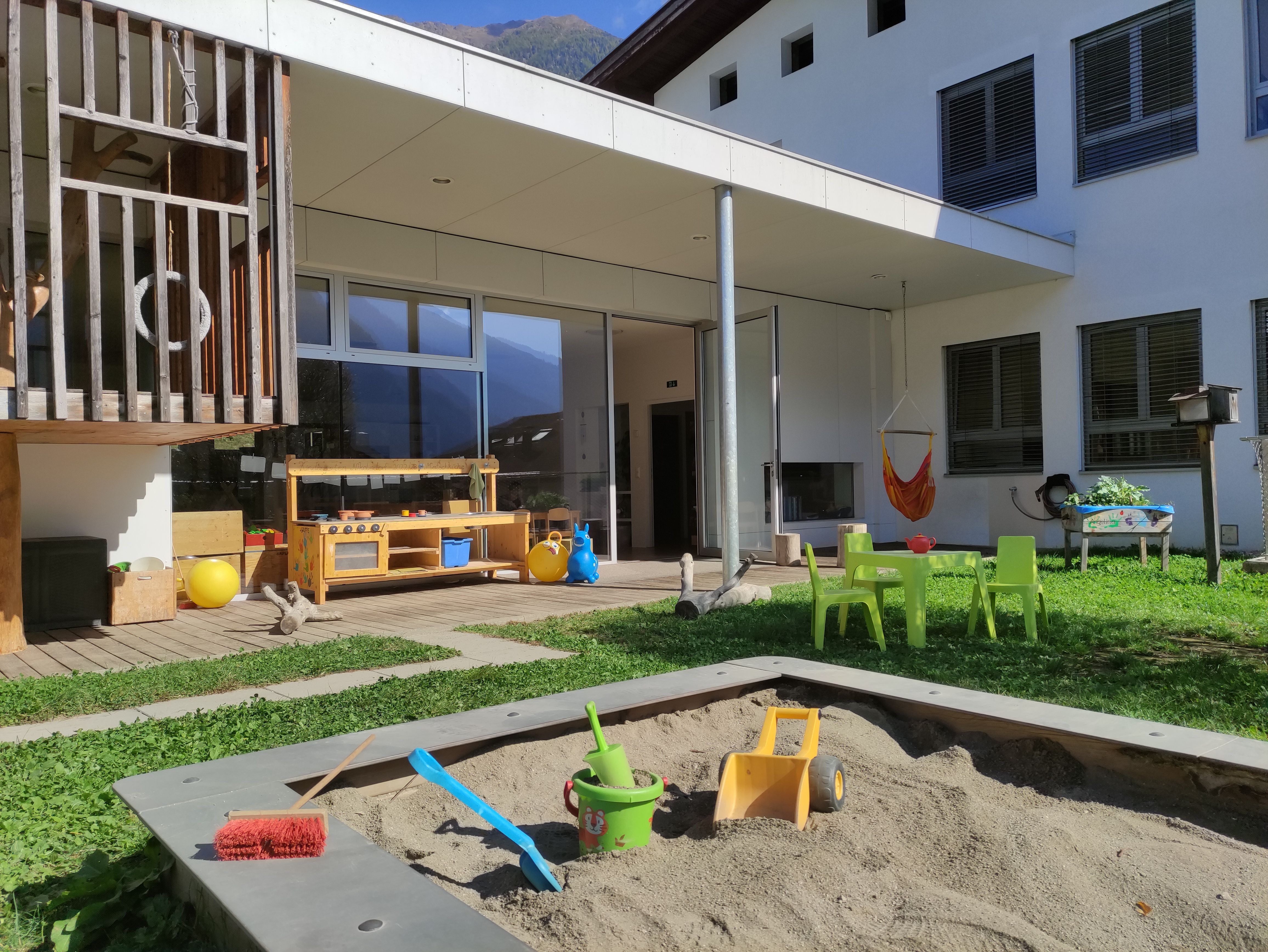 Zusehen ist der Außenbereich des Kindergarten. Im Vordergrund ist ein Sandkaten mit bunten Spielsachen. Dahinter stehen grüne Kinderstühle und ein Tisch auf einer Rasenfläche.